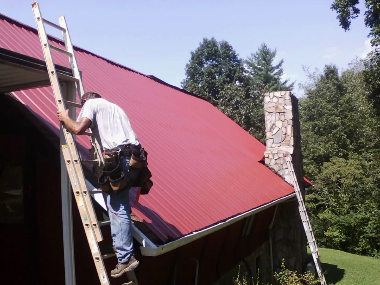 Roofer on a ladder beside a red metal roof and stone chimney — Soffit & Fascia Repair by Roofing Brothers in Brooklyn